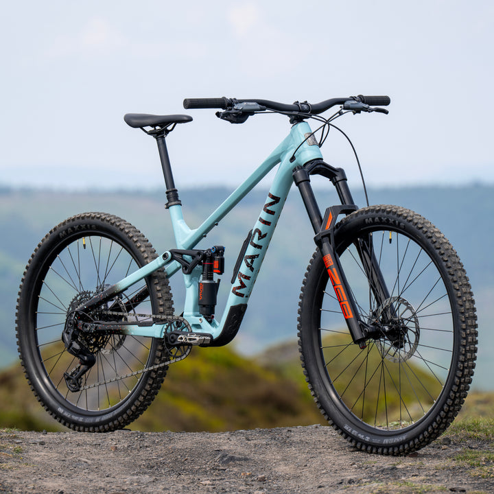 A Marin Alpine Trail 2 full-suspension enduro mountain bike in ash blue, parked on a rocky outcrop with a blurred natural landscape in the background.